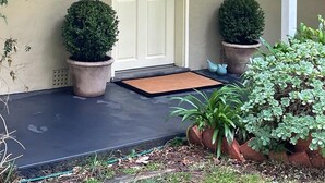 Une maison avec une porte blanche, deux arbustes taillés en forme décorative placés dans des pots, un paillasson et un petit jardin.