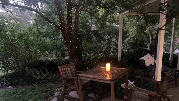 Une terrasse extérieure avec une table et des chaises en bois, entourée de chemins de pierre et d’une végétation luxuriante.