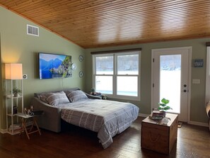 Une chambre à coucher comprenant un lit, une table de chevet, une lampe, un téléviseur et une fenêtre donnant sur un lac.