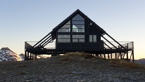 A modern mountain lodge with a triangular roof and large windows, situated on a rocky hillside with a view of snow-capped mountains. - Exclusive Alpine Chalet overlooking Mt Aspiring National Park (Wanaka)