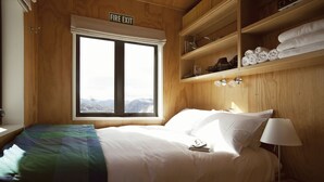 A hotel room with a bed, a nightstand, a lamp, a shelf with towels, and a window with a view of mountains. - Exclusive Alpine Chalet overlooking Mt Aspiring National Park (Wanaka)