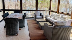 A covered outdoor seating area with wicker furniture, a table, and a view of a forest. - Luxury Lake Lanier Estate + Double Dock (Gainesville)