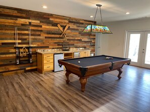 A pool table in a room with a wooden floor and a kitchen area.