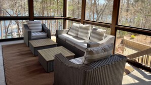 A covered outdoor seating area with wicker furniture and large windows offering a view of a forest and a body of water. - Luxury Lake Lanier Estate + Double Dock (Gainesville)
