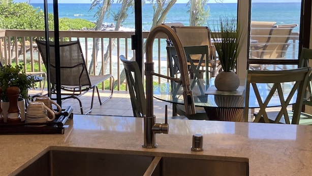 Una cocina con un lavamanos, ventana con vista al océano y mobiliario exterior con sillas y una mesa.