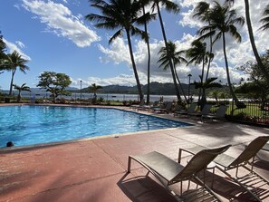 A swimming pool with lounge chairs and palm trees, with a view of the ocean and mountains in the background.