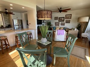 A modern kitchen with a central island, dining area with green chairs, and a living room with a sofa and decorative pillows.
