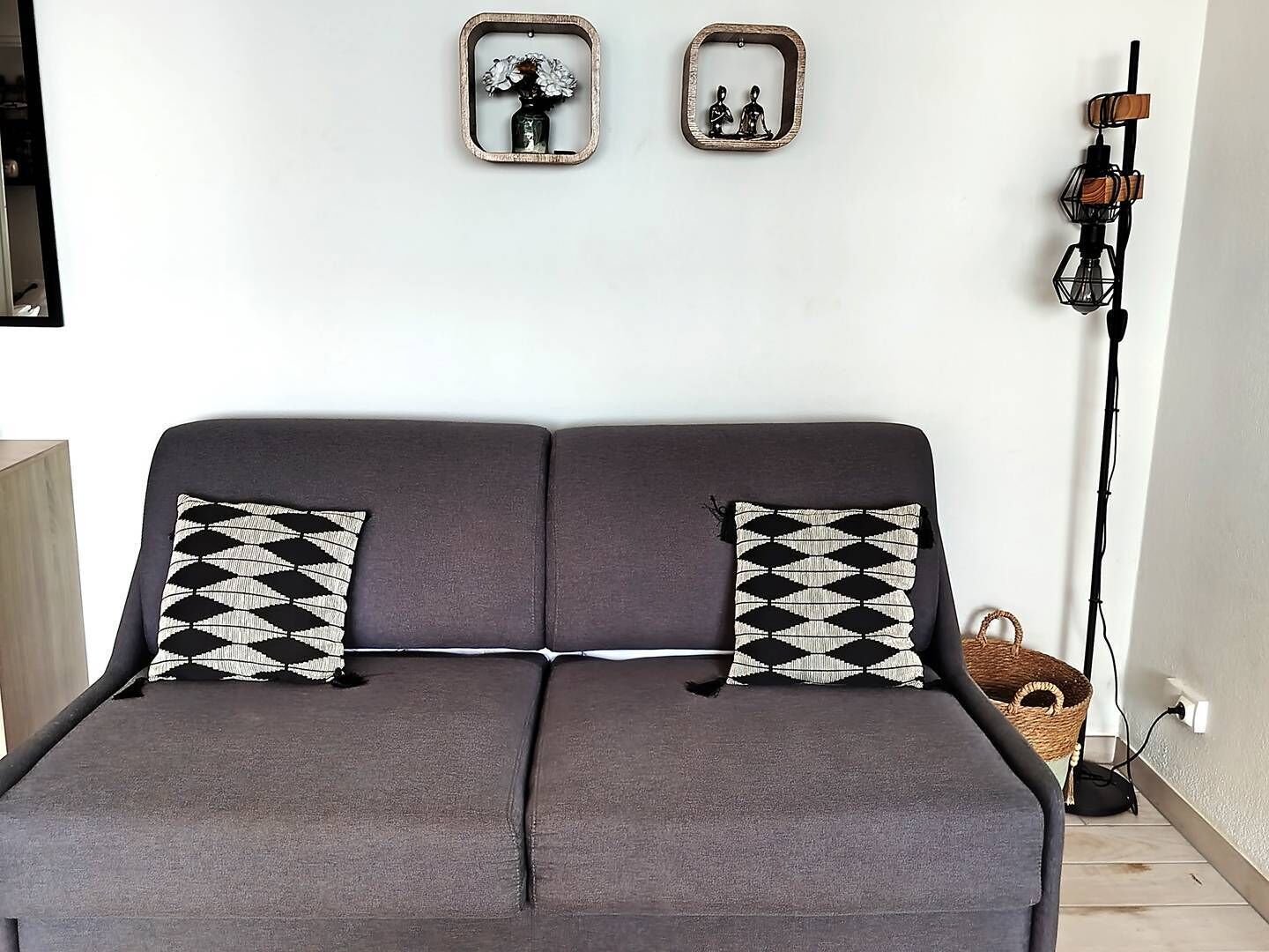 A grey sofa with two patterned pillows, a wall-mounted light fixture, and two framed pictures above it.