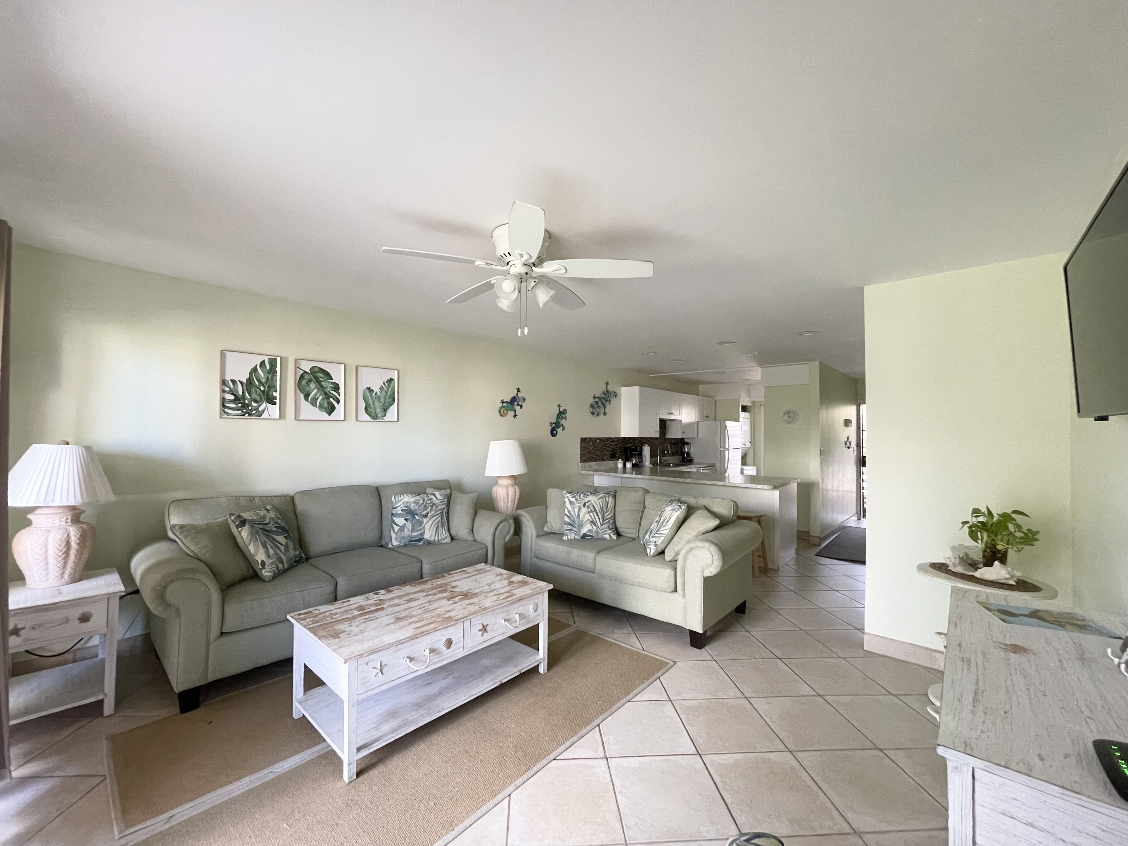 A living room with a sofa, coffee table, and a ceiling fan. There is a kitchen in the background.