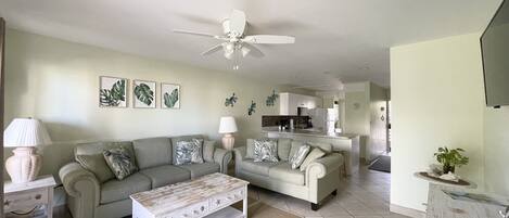 A living room with a sofa, coffee table, and a ceiling fan. There is a kitchen in the background.