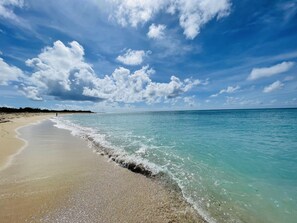Ein Strand mit klarem türkisblauem Wasser, einem Sandufer und einem teilweise bewölkten Himmel.