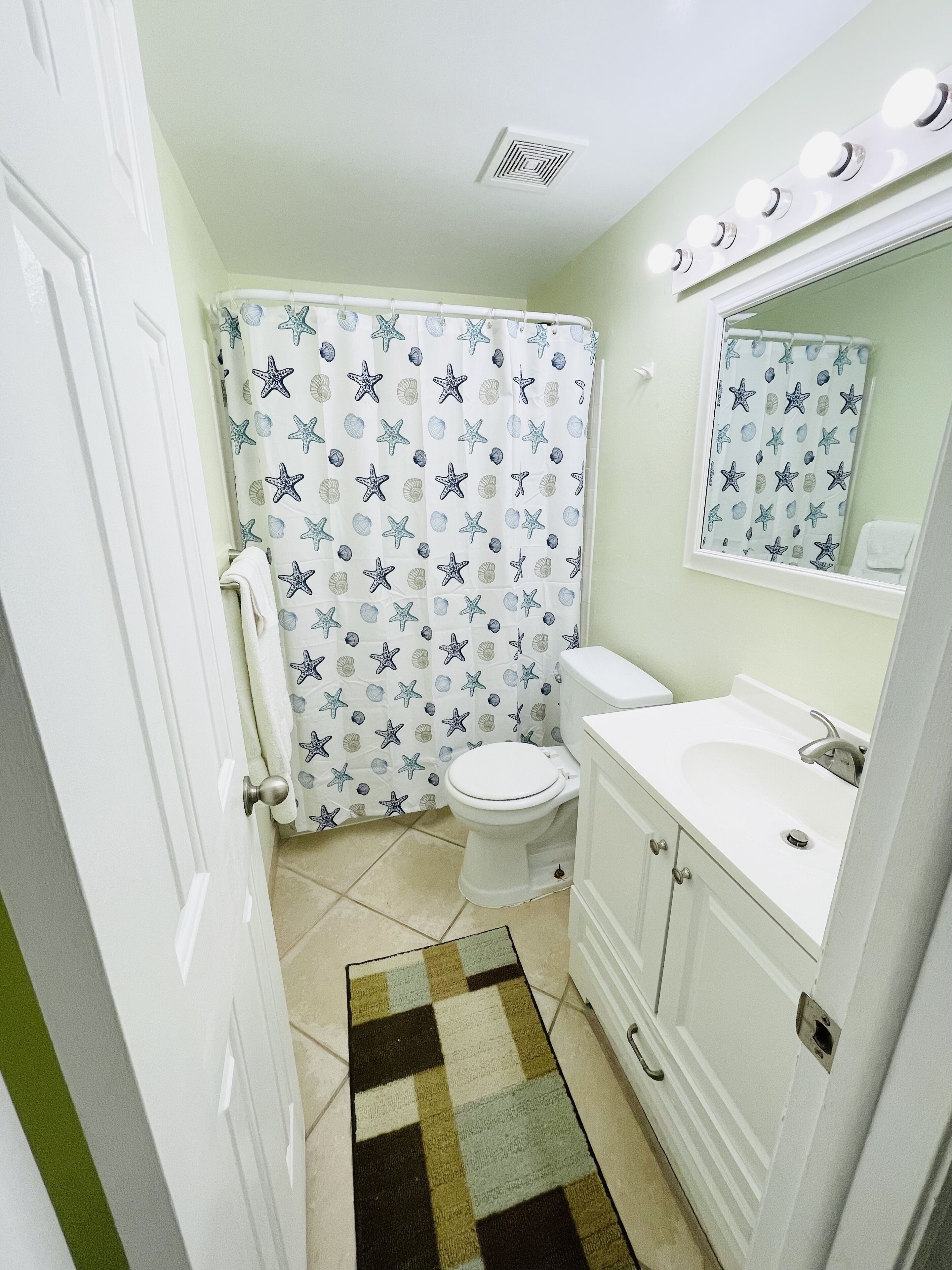 Baño con inodoro, lavamanos, espejo y cortina de ducha con estrellas de mar y conchas marinas.