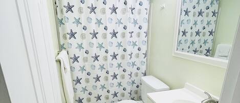 Baño con inodoro, lavamanos, espejo y cortina de ducha con estrellas de mar y conchas marinas.