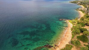 Vue aérienne d’une zone côtière avec des eaux turquoise limpides, une plage de sable et un paysage montagneux en arrière-plan.
