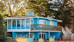 Une maison bleue avec un balcon, entourée de chaises de plage et d’une zone sablonneuse.