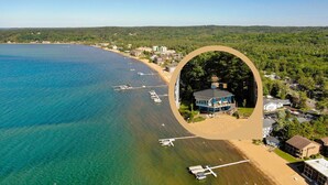 Vue aérienne d’une zone côtière avec une plage, une eau d’un bleu limpide et un petit bâtiment entouré de verdure.