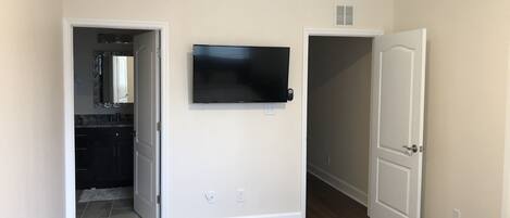 Habitació amb televisió de pantalla plana penjada a la pared, una porta que condueix a una altra habitació i un bany visible amb mirall i rentapins.