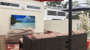 A covered outdoor seating area with a wicker sofa, a red umbrella, and a television displaying a beach scene. - Newly constructed modern home with luxurious rooftop patio (Tallahassee)
