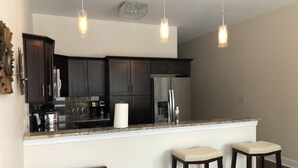 A modern kitchen with dark cabinets, a stainless steel refrigerator, a marble countertop, and three bar stools. - Newly constructed modern home with luxurious rooftop patio (Tallahassee)