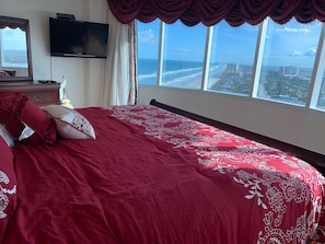 A bedroom with a large bed, a TV mounted on the wall, and a view of the ocean and cityscape through large windows.