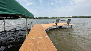 Eine Holzstele mit zwei Stühlen und einem Tisch, ein grünes Blattenzelt und ruhiges Wasser.