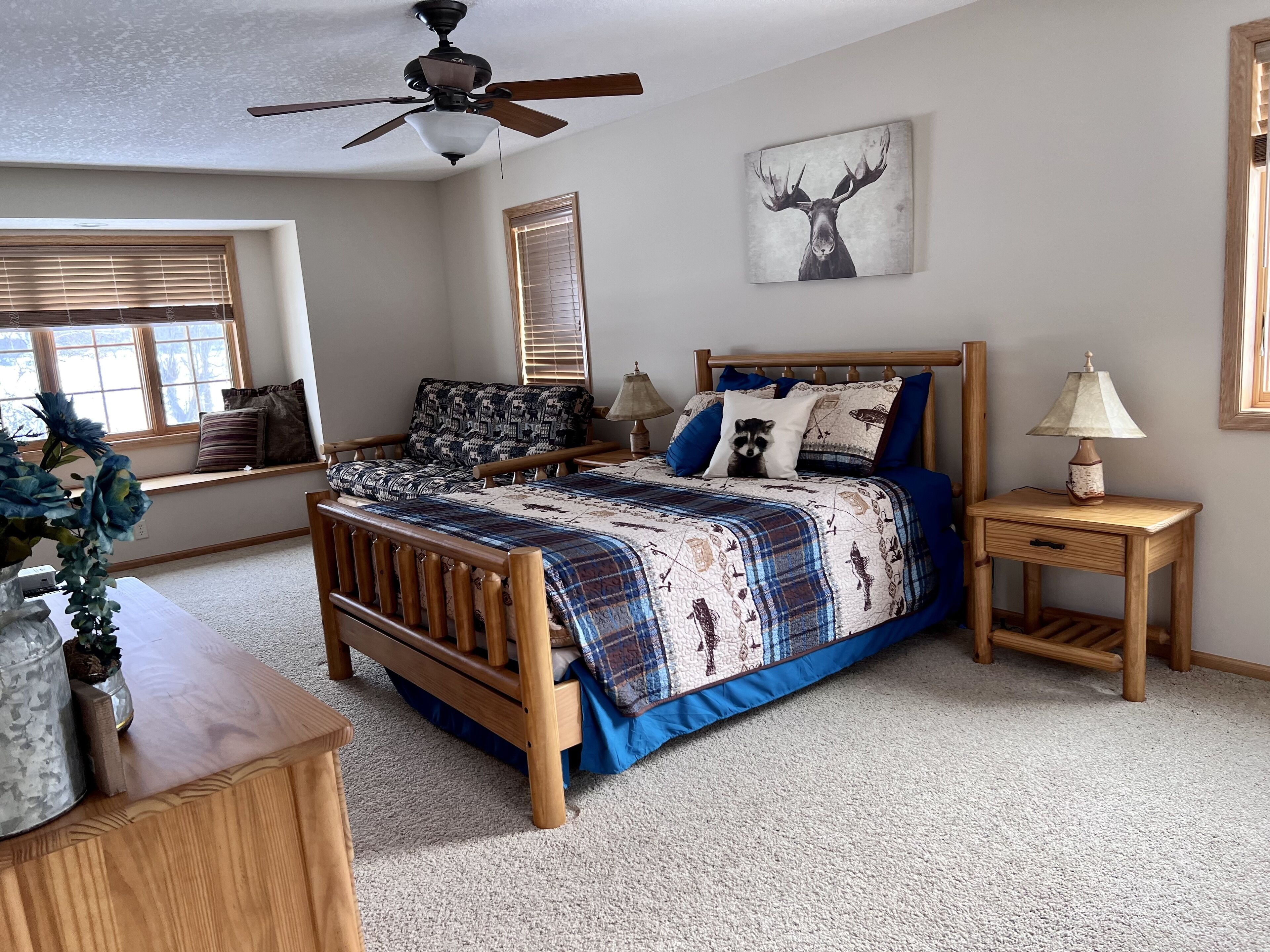 A bedroom with a wooden bed, a ceiling fan, a window with blinds, a wall-mounted lamp, and a raccoon-themed pillow.