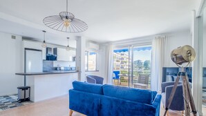 A modern living room with a blue sofa, a dining area with a kitchen, and a balcony with a view of buildings. - 2Br. Apartment, 100m from Beaches, Terrace & Parking (Cannes)