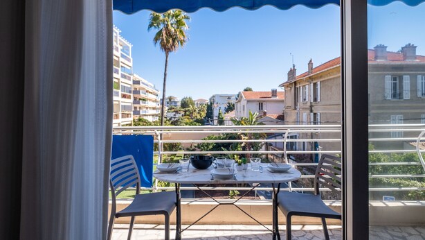 A balcony with a table set for dining, overlooking a residential area with buildings and a palm tree. - 2Br. Apartment, 100m from Beaches, Terrace & Parking (Cannes)