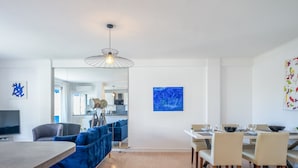 A modern living room with a blue sofa, a dining area with a white table, and a kitchen in the background. - 2Br. Apartment, 100m from Beaches, Terrace & Parking (Cannes)