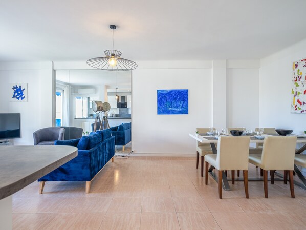 Una sala de estar moderna con un sofá azul, un comedor con una mesa blanca y una cocina al fondo.