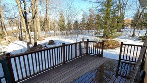 A wooden deck with a railing, surrounded by snow-covered ground and trees.
