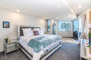 A modern bedroom with a large bed, a glass side table, a mirror, and a view of the outdoors.