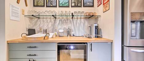 Cozinha equipada com máquina de lavar louça, cafeteira e frigorífico. Quadros emoldurados na parede e uma estante com copos e chávenas.