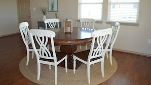 Un coin repas avec une table ronde en bois entourée de chaises blanches, un tapis tissé et une porte visible au fond.