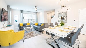 Un salon moderne avec un coin repas, des canapés gris agrémentés de coussins jaunes, un fauteuil jaune, une grande table à manger avec des chaises et un ventilateur de plafond.