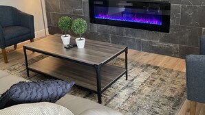 A modern living room with a stone wall, a flat-screen TV mounted above a fireplace, a coffee table with potted plants, and a sofa with a throw pillow. - Comfortable home with sparkling private pool minutes to the Strip! (Henderson)