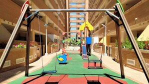 Zona de juegos infantiles con columpios y un tobogán, rodeada de muros de ladrillo y macetas con plantas.