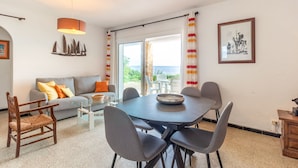 A modern living room with a grey sofa, a glass coffee table, and a dining table with grey chairs. There is a large window with a view of the sea, a hanging light fixture, and a framed picture on the wall. - Carboneras 48 - Apartment on the seafront (Llança)