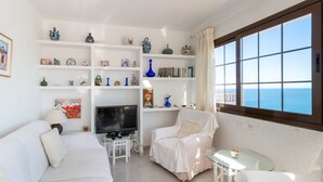 Ein Wohnzimmer mit einer Couch, einem Fernseher, einem Glastisch und einem Sessel. Regale mit Dekorationen und Blick auf das Meer durch ein Fenster.