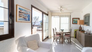 Ein helles Wohnzimmer mit einem weißen Sofa, einem Esstisch mit Stühlen, einem Deckenventilator und Meerblick durch große Fenster.