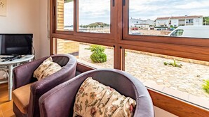 Una habitación con un gran ventanal, dos sillones de cuero con cojines estampados, una mesita con un control remoto y un televisor sobre un mueble.