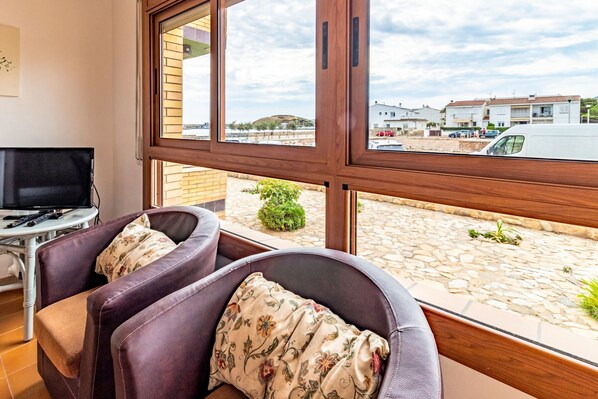 A room with a large window, two leather armchairs with floral cushions, a small table with a remote control, and a TV on a stand.