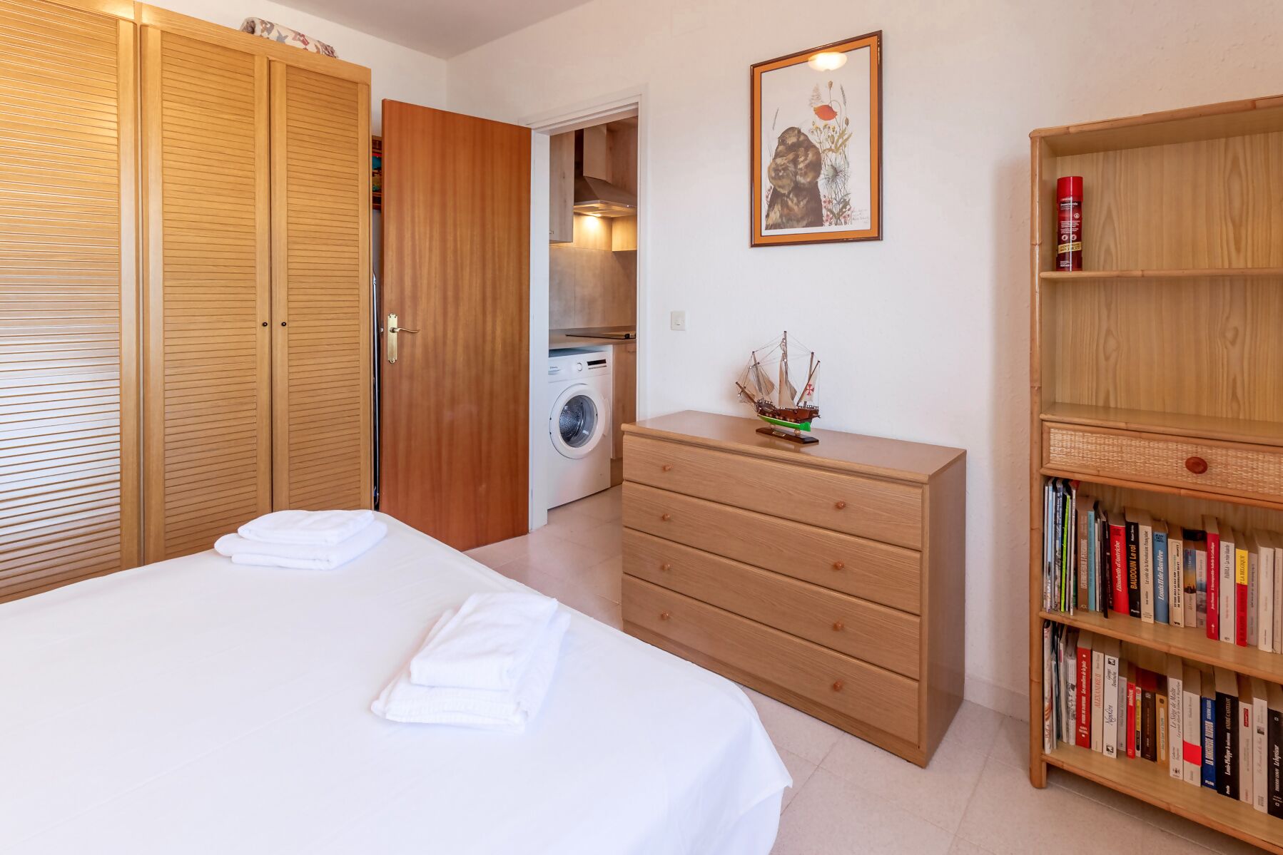 Ein Schlafzimmer mit Bett, Holzschrank, Kommode, Bücherregal und einer Waschmaschine im Hintergrund.