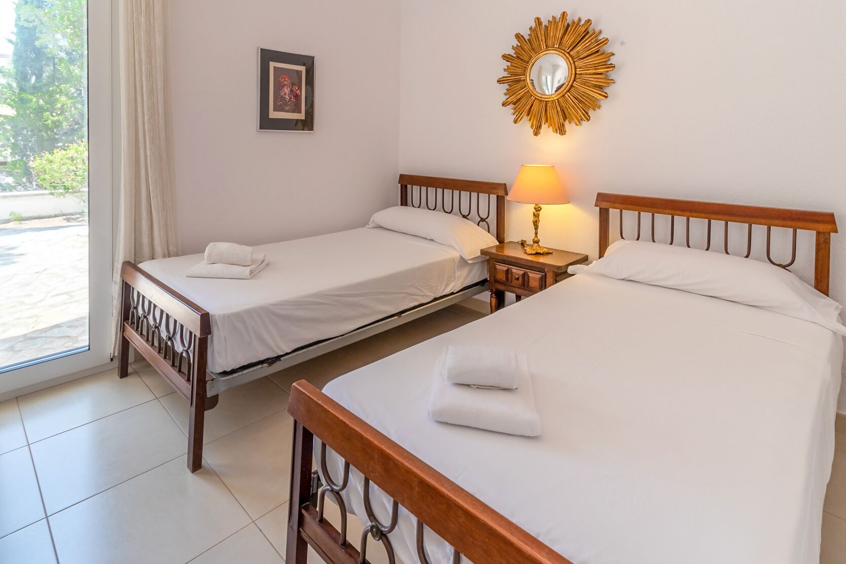 Habitación de hotel con dos camas individuales, una mesita de noche con lámpara, un cuadro en la pared y vista al exterior a través de una ventana.