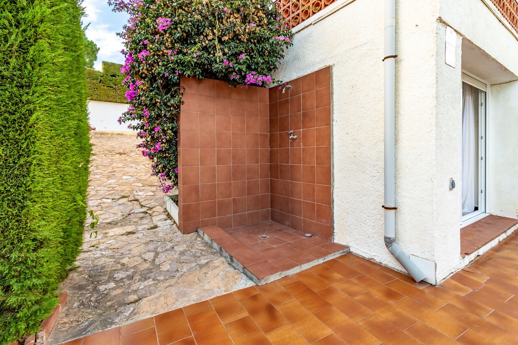 Un área de ducha exterior pequeña y cerrada, con paredes y suelo de baldosas, junto a un edificio blanco con una puerta y una ventana.
