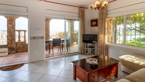 Un soggiorno spazioso con divano, tavolino e TV. Presente un balcone con colonna in pietra e un'area pranzo con sedie.