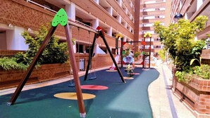 Ein Kinderspielbereich mit Schaukeln und einem farbenfrohen Boden, umgeben von Ziegelmauern und Grünpflanzen.