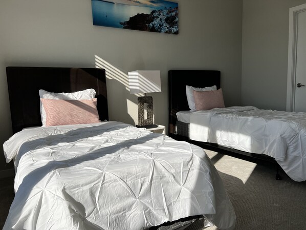 Ein Hotelzimmer mit zwei Betten, jeweils mit weißer Bettwäsche und Kissen mit rosa Muster. Dazwischen befindet sich ein Nachttisch mit einer Lampe. Über den Betten hängt ein Gemälde, das eine Küstenlandschaft zeigt.