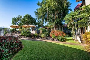 A well-maintained garden with a lawn, various shrubs, and trees. There are outdoor seating areas with umbrellas. - Walk to Charming Kailua Village. Kailua Bay View. Lanai. AC. King Bed. Pool. (Kailua-Kona)