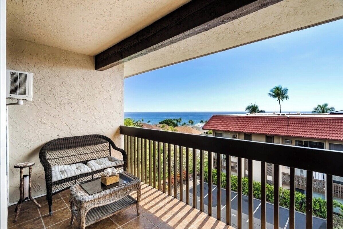 A balcony with a wicker sofa and table, overlooking a coastal view with buildings and palm trees.